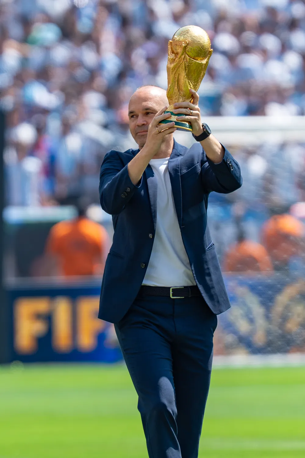 짙은 남색 정장을 입고 FIFA 월드컵 트로피를 들어 보이며 경기장으로 걸어 들어오는 안드레스 이니에스타