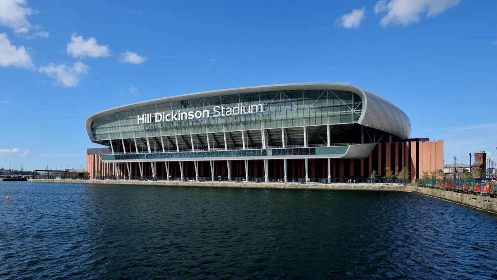 푸른 물가 위로 은빛 곡선 지붕과 Hill Dickinson Stadium 글자가 보이는 에버턴 새 경기장