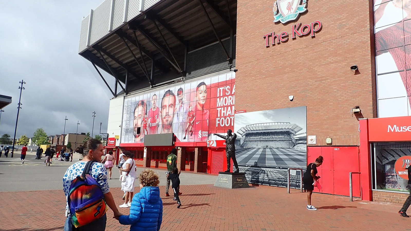 THE KOP 글자와 리버풀 문장이 걸린 안필드 콥 스탠드 외관과 그 앞을 걷는 가족