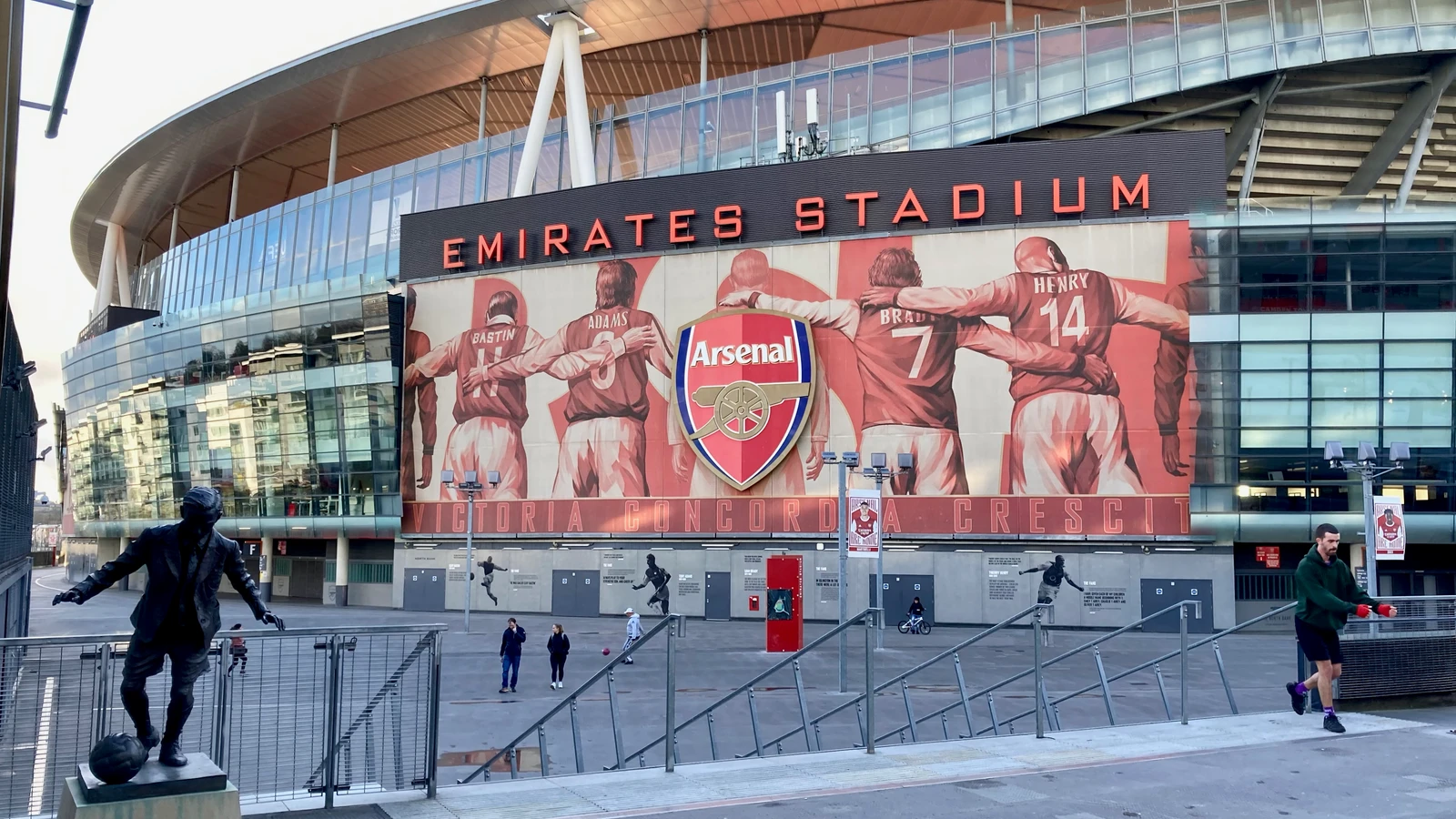아스널 선수들이 어깨동무한 벽화와 EMIRATES STADIUM 글자가 보이는 경기장 외관과 동상