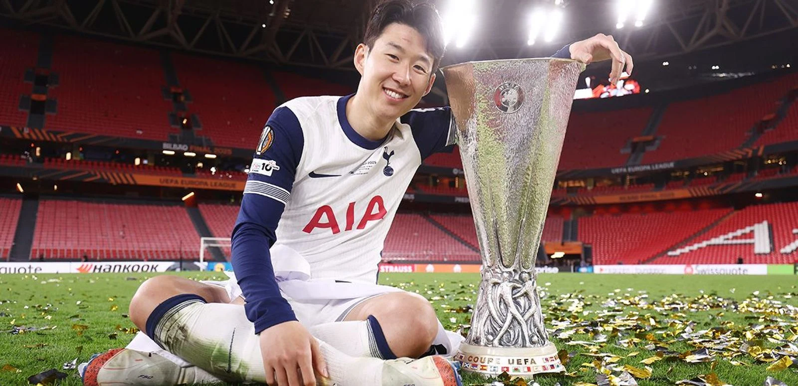 텅 빈 산 마메스 경기장 잔디 위에 앉아 UEFA 유로파 리그 트로피에 손을 얹고 웃는 손흥민